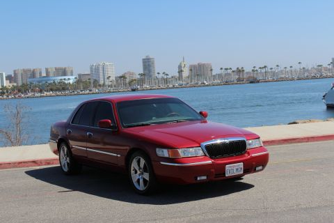 2002 Mercury Grand Marquis - A red 2002 Mercury Grand Marquis is parked near a waterfront with a city skyline in the distance.