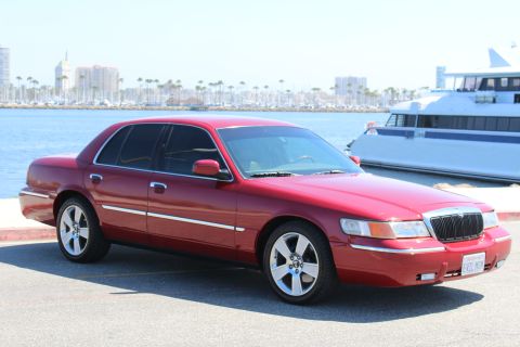2002 Mercury Grand Marquis - A red 2002 Mercury Grand Marquis is parked near the water with a large white boat in the background.
