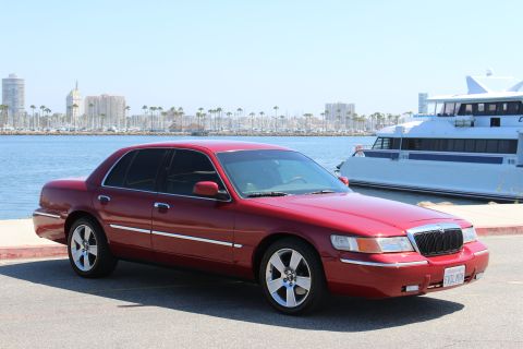 2002 Mercury Grand Marquis - A red 2002 Mercury Grand Marquis is parked near a harbor with a large white boat and city skyline visible.