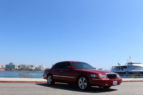 2002 Mercury Grand Marquis - A red 2002 Mercury Grand Marquis is parked near a waterfront with a large passenger ferry in the background.