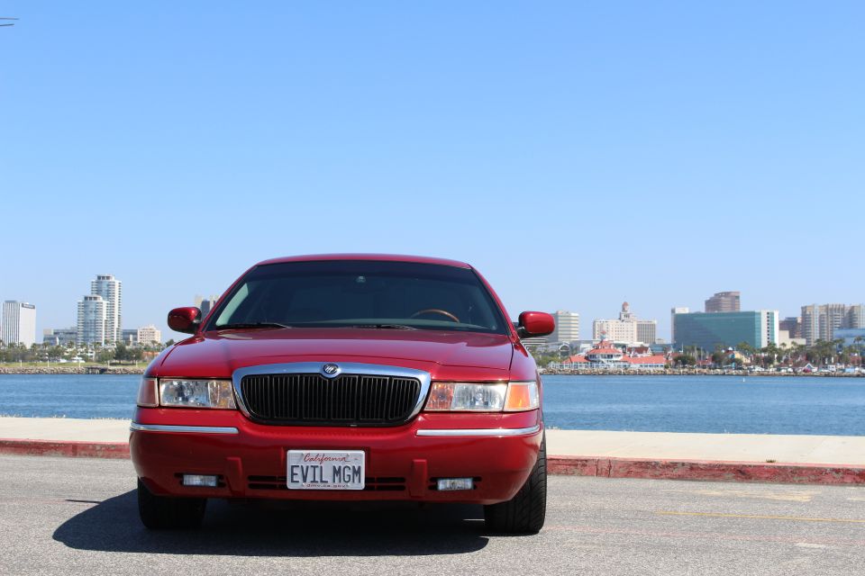 Red 2002 Mercury Grand Marquis parked by the water with a city skyline in the background. License plate reads EVIL MGM