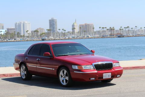 2002 Mercury Grand Marquis - A red 2002 Mercury Grand Marquis is parked near a harbor with a city skyline visible.