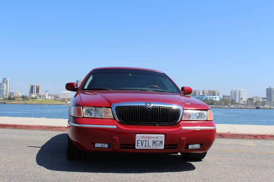 Red 2002 Mercury Grand Marquis parked by the water with city skyline in background. License plate says EVIL MGM