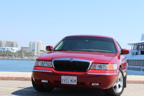 2002 Mercury Grand Marquis - A red 2002 Mercury Grand Marquis is parked by the water with buildings in the background.