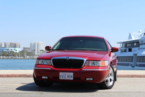 2002 Mercury Grand Marquis - A red 2002 Mercury Grand Marquis is seen from the front, parked near a body of water with a large boat and city skyline behind it.