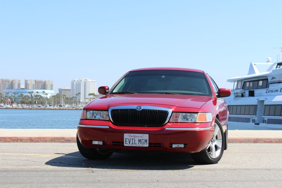 2002 Mercury Grand Marquis front view, parked near water with a boat and city buildings in background. License plate reads EVIL MGM