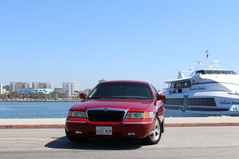 2002 Mercury Grand Marquis - A red 2002 Mercury Grand Marquis is parked in front of a large passenger ferry with a city skyline in the background.