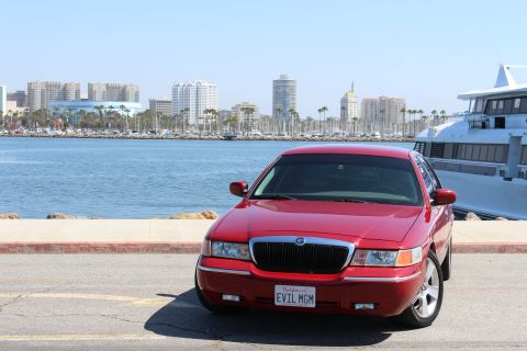 2002 Mercury Grand Marquis - A red 2002 Mercury Grand Marquis is parked near a waterfront with a city skyline and a large yacht in the background.