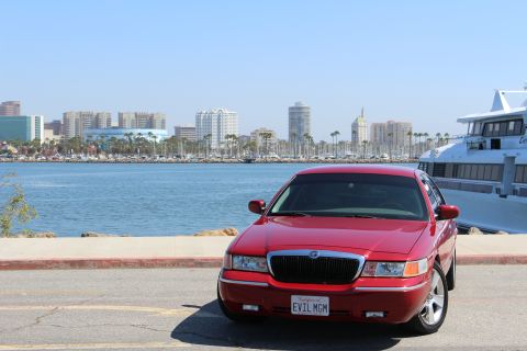 2002 Mercury Grand Marquis - A red 2002 Mercury Grand Marquis is parked near a harbor with a city skyline and a large yacht.