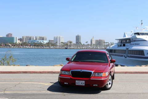 2002 Mercury Grand Marquis - A red 2002 Mercury Grand Marquis is parked near a harbor with a city skyline and a large passenger ferry visible.