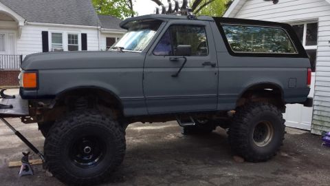 1994 Ford Bronco - Driver-side profile of a gray primer-finished full-size Ford Bronco with a black removable hardtop, lifted on mud-terrain tires and black s…