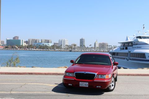 2002 Mercury Grand Marquis - A red 2002 Mercury Grand Marquis is parked near a waterfront with a city skyline and a large boat in the background.