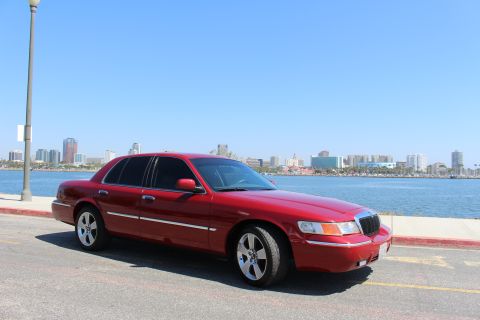 2002 Mercury Grand Marquis - A red 2002 Mercury Grand Marquis is parked near a body of water with a city skyline visible in the distance.