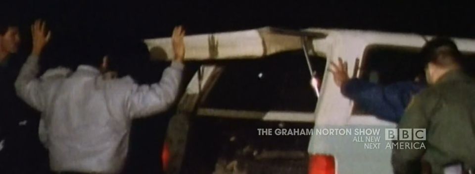 People with hands up near the rear of a dark SUV with a white roof rack
