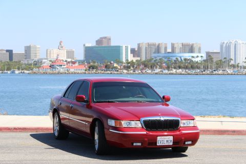 2002 Mercury Grand Marquis - A red 2002 Mercury Grand Marquis is parked by the water with a city skyline in the background.