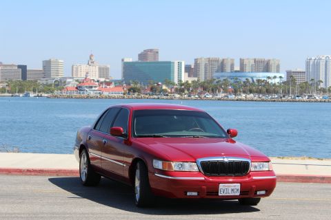 2002 Mercury Grand Marquis - A red 2002 Mercury Grand Marquis is parked near a body of water, with a city skyline visible behind it.