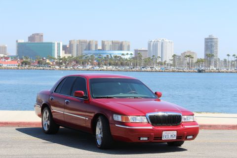 2002 Mercury Grand Marquis - A red 2002 Mercury Grand Marquis is parked near a waterfront with a city skyline in the distance.