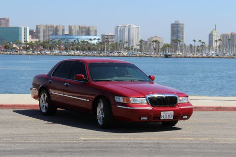 2002 Mercury Grand Marquis - A red 2002 Mercury Grand Marquis is parked near a body of water with a city skyline in the background.