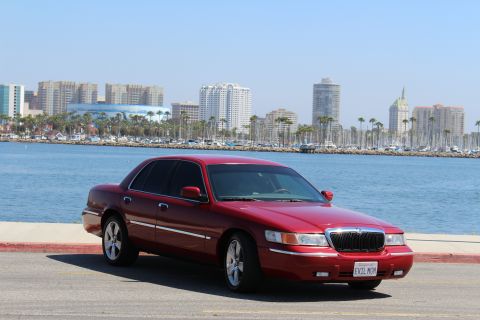 2002 Mercury Grand Marquis - A red 2002 Mercury Grand Marquis is parked near a waterfront with a city skyline and boats in the background.