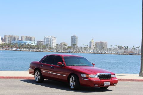 2002 Mercury Grand Marquis - A red 2002 Mercury Grand Marquis is parked by the water with a city skyline and palm trees in the background.