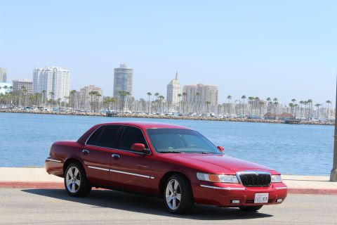 2002 Mercury Grand Marquis - A red 2002 Mercury Grand Marquis is parked near a waterfront with a city skyline in the background.