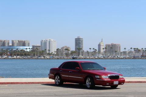 2002 Mercury Grand Marquis - A red 2002 Mercury Grand Marquis is parked by the water with a city skyline and palm trees in the distance.