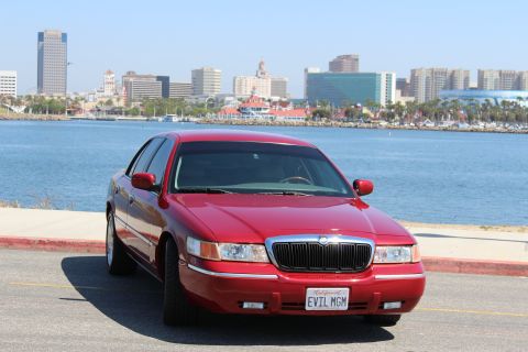 2002 Mercury Grand Marquis - A red 2002 Mercury Grand Marquis is parked near a body of water with a city skyline in the distance.