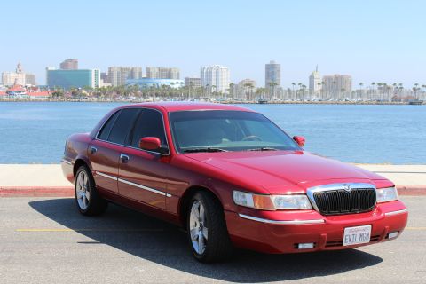 2002 Mercury Grand Marquis - A red 2002 Mercury Grand Marquis is parked near a body of water with a city skyline visible in the distance.