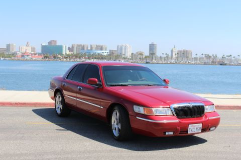 2002 Mercury Grand Marquis - A red 2002 Mercury Grand Marquis is parked near a body of water with a city skyline in the distance.