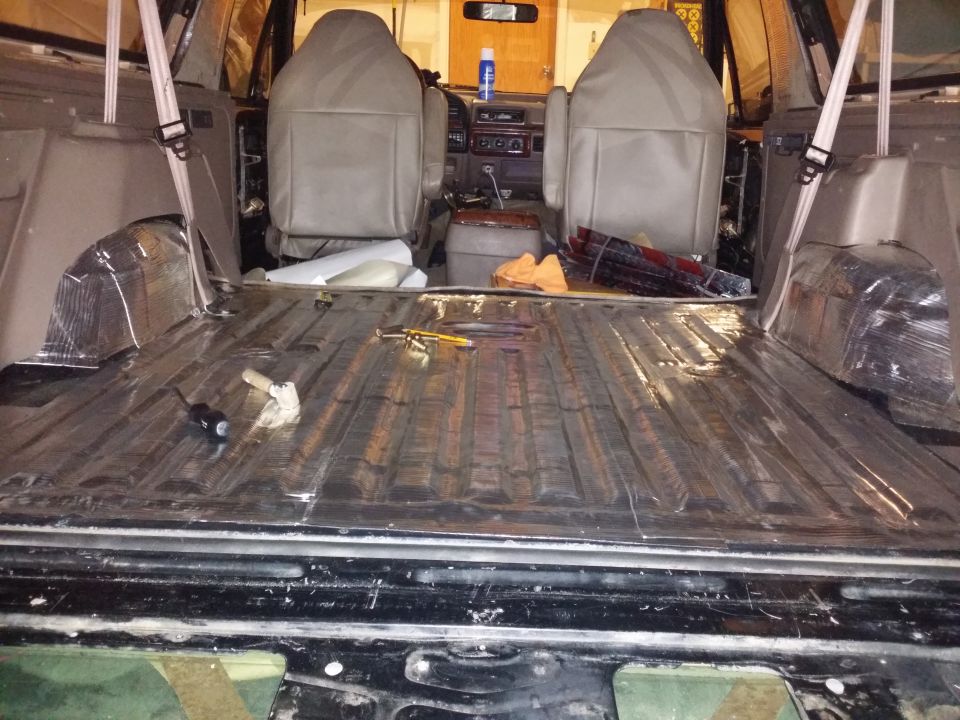 Interior of a vehicle with seats removed, showing soundproofing material on the floor and wheel wells. Tools are on the floor