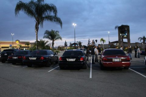 2003 Ford Crown Victoria - Several sedans, mostly black with one maroon, parked rear-on at dusk in a strip-mall lot near a McDonald's sign.