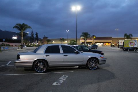 2003 Ford Crown Victoria - Silver Ford sedan on large chrome multi-spoke wheels parked side-on in a strip-mall lot at dusk near a Lay's billboard.