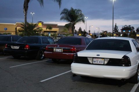 2003 Ford Crown Victoria - white Ford bearing a WHTE VIC plate, a maroon Mercury, and a black sedan sit angled in a strip-mall lot at dusk.