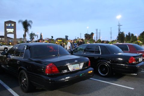2003 Ford Crown Victoria - Two black Ford full-size sedans on dark wheels park rear-first while a small crowd gathers in a shopping center lot at dusk.