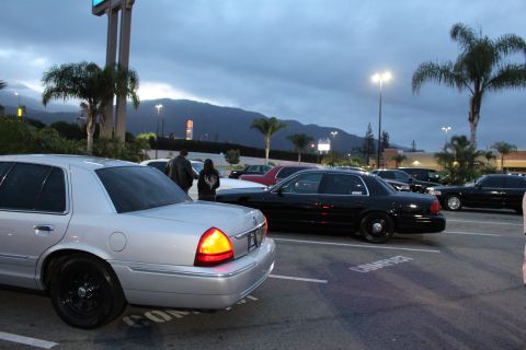 2003 Ford Crown Victoria - silver Ford sedan and a black full-size sedan park in a lot at dusk with palm trees and a dark mountain ridge behind.