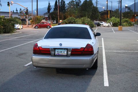 2003 Ford Crown Victoria - rear of a white Mercury Grand Marquis with a MAH MERC vanity plate sits in a near-empty lot, a red VW Beetle behind it.