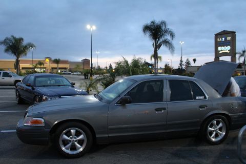 2003 Ford Crown Victoria - gray Ford Crown Victoria with its hood raised parks among other full-size sedans in a shopping center lot at dusk.
