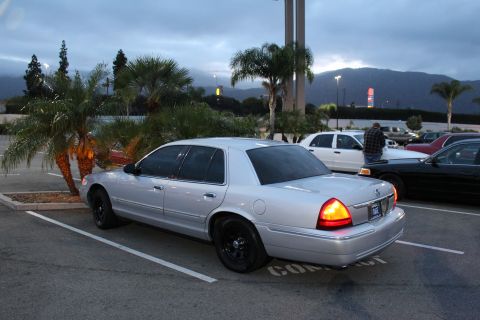 2003 Ford Crown Victoria - silver Ford Crown Victoria on black wheels parks beside palm trees in a lot with a mountain ridge and overcast sky at dusk.