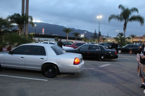 2003 Ford Crown Victoria - silver Ford sedan and black sedans park in a strip-mall lot at dusk as people stand nearby beneath palm trees and mountains.