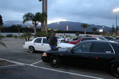 2003 Ford Crown Victoria - black Crown Victoria and a white Ford sedan flank a man walking between rows at a dusk car meet with mountains behind.