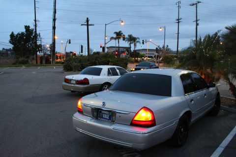 2003 Ford Crown Victoria - silver Mercury sedan with glowing tail lights and a beige sedan sit near a lit road intersection at dusk under power lines.