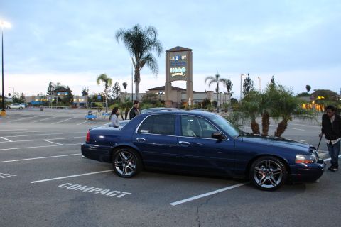 2003 Ford Crown Victoria - dark blue Mercury Grand Marquis on chrome wheels parks in profile in a strip-mall lot as people stand near it at dusk.
