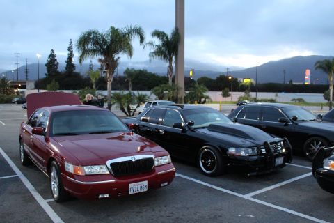 2003 Ford Crown Victoria - maroon Mercury with an EVIL MOM plate parks beside a black Crown Victoria fitted with a push bar in a lot at dusk.