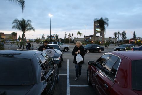 2003 Ford Crown Victoria - blonde woman carrying a white bag walks between parked cars while a man holds a shirt at a dusk parking-lot meet.