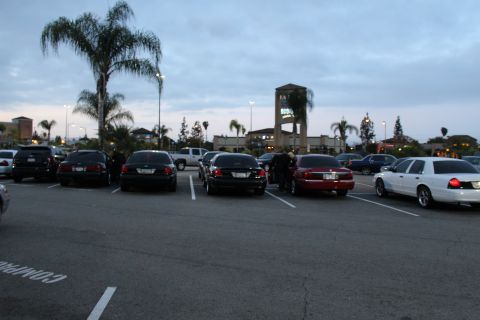 2003 Ford Crown Victoria - row of dark sedans and a white Ford sit across a wide shopping center parking lot near a lit clock tower at dusk.