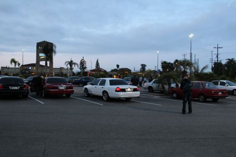2003 Ford Crown Victoria - white Ford and other cars are scattered across a wide parking lot as people stand among them near a clock tower at dusk.