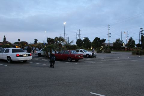 2003 Ford Crown Victoria - white Ford, a red compact sedan and other cars sit in a wide open parking lot with people walking at twilight.