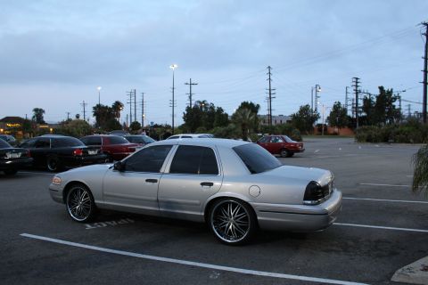 2003 Ford Crown Victoria - silver full-size sedan on large chrome multi-spoke wheels sits in an empty parking lot at dusk with other cars and palm trees behind.