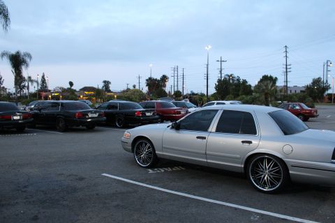 2003 Ford Crown Victoria - silver full-size sedan on large chrome wheels parked in a lot at dusk, with a row of dark sedans and palm trees in the background.