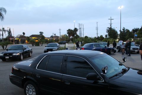 2003 Ford Crown Victoria - black full-size sedan with heavily tinted windows parked in a lot at dusk, with other cars and a few people gathered behind.